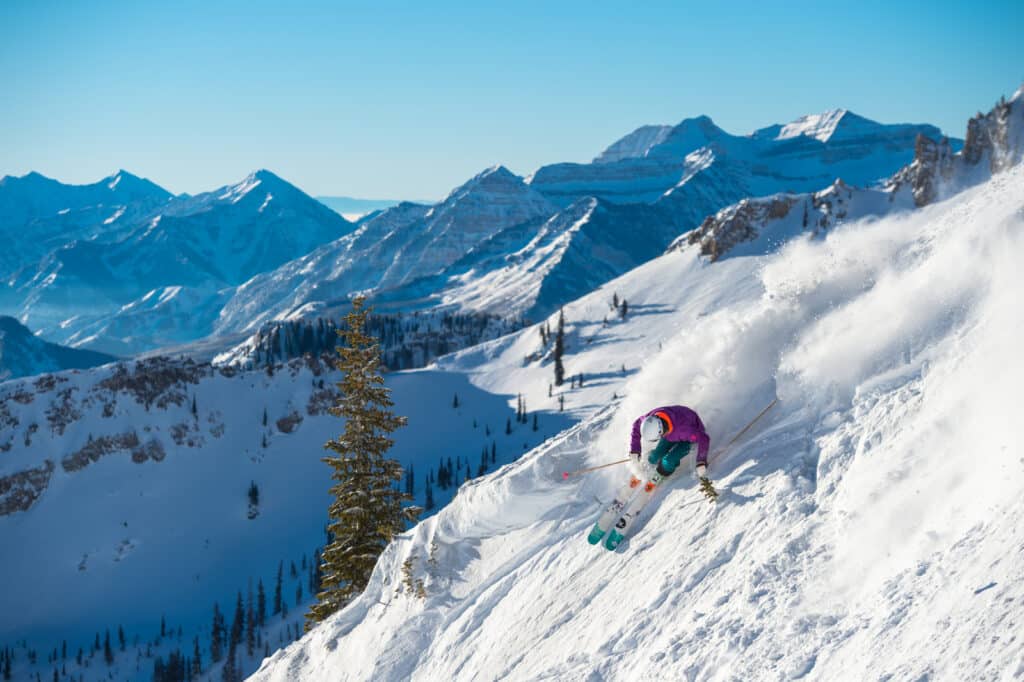 Katie Van Riper skiing through powder down a steep slope with a view of mountain peaks behind her on a bluebird day at Snowbird Ski Resort up Little Cottonwood Canyon, Utah